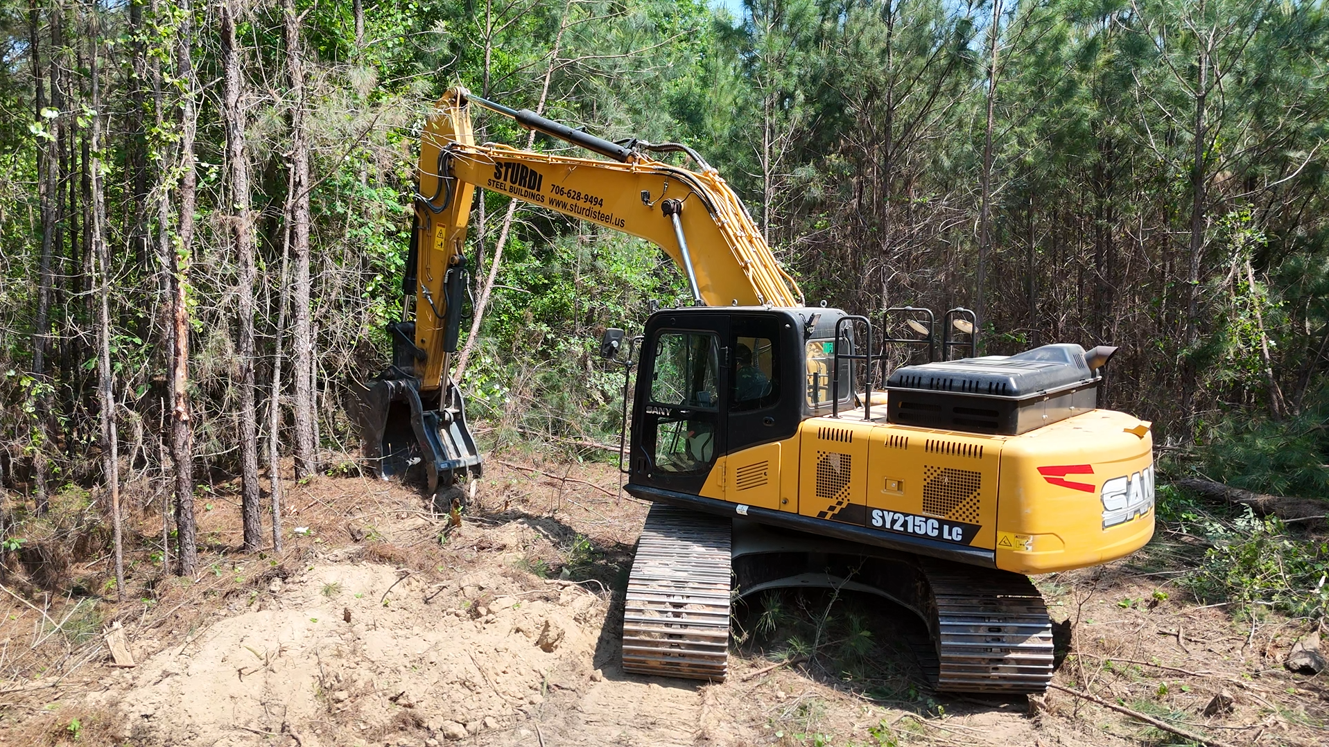 Yellow excavator clearing a narrow dirt path beside dense trees in a wooded area