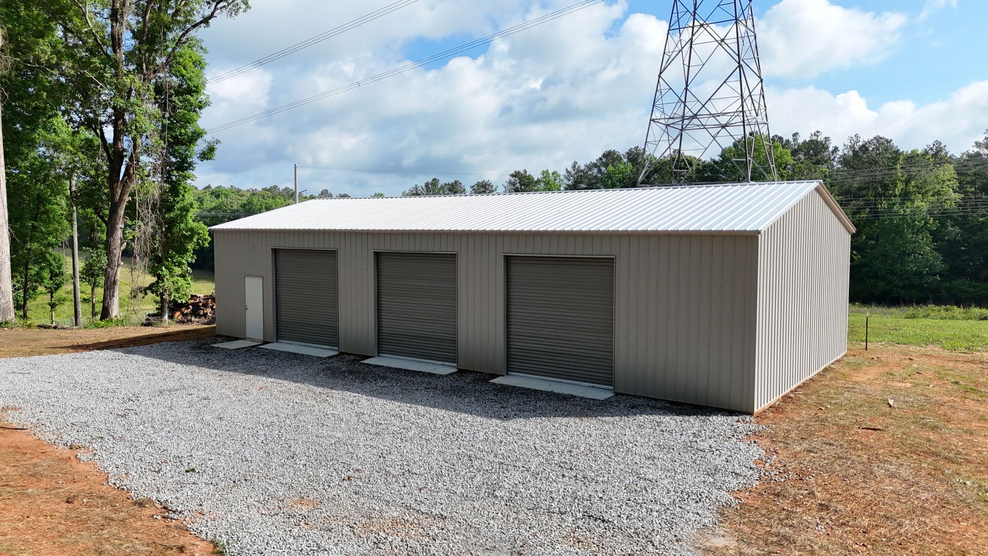 A metal building with three garage doors is sitting in the middle of a gravel driveway.