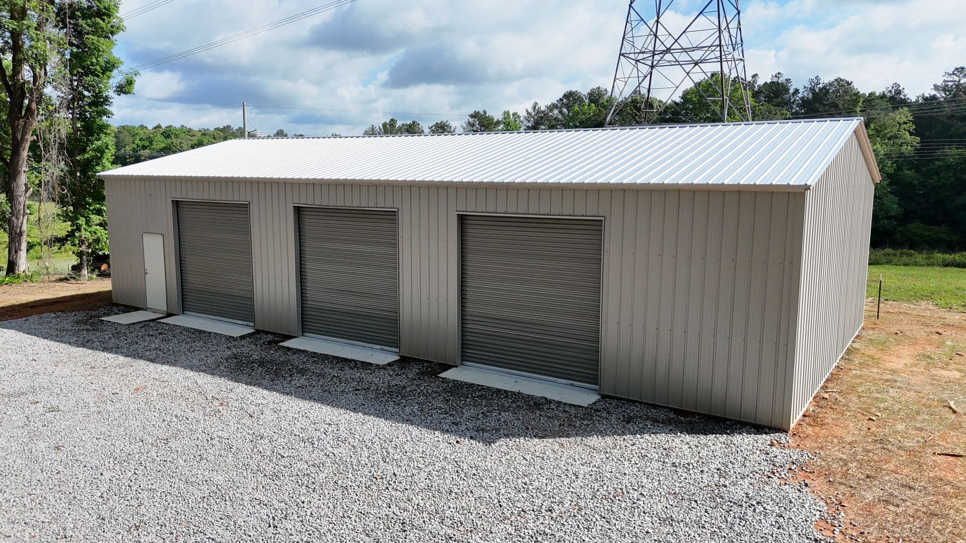 A large metal building with three garage doors is sitting on top of a gravel lot.