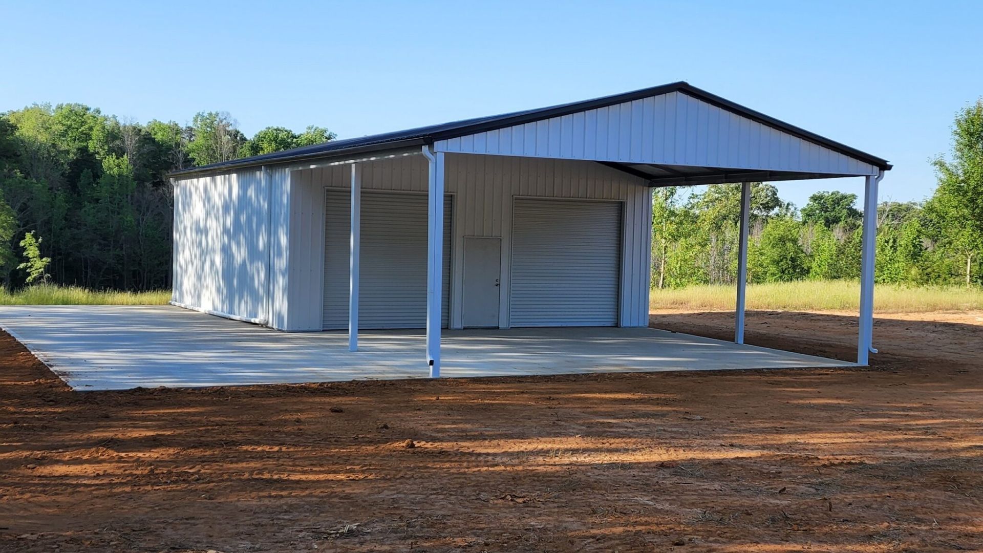 A white garage with a black roof is sitting in the middle of a dirt field.