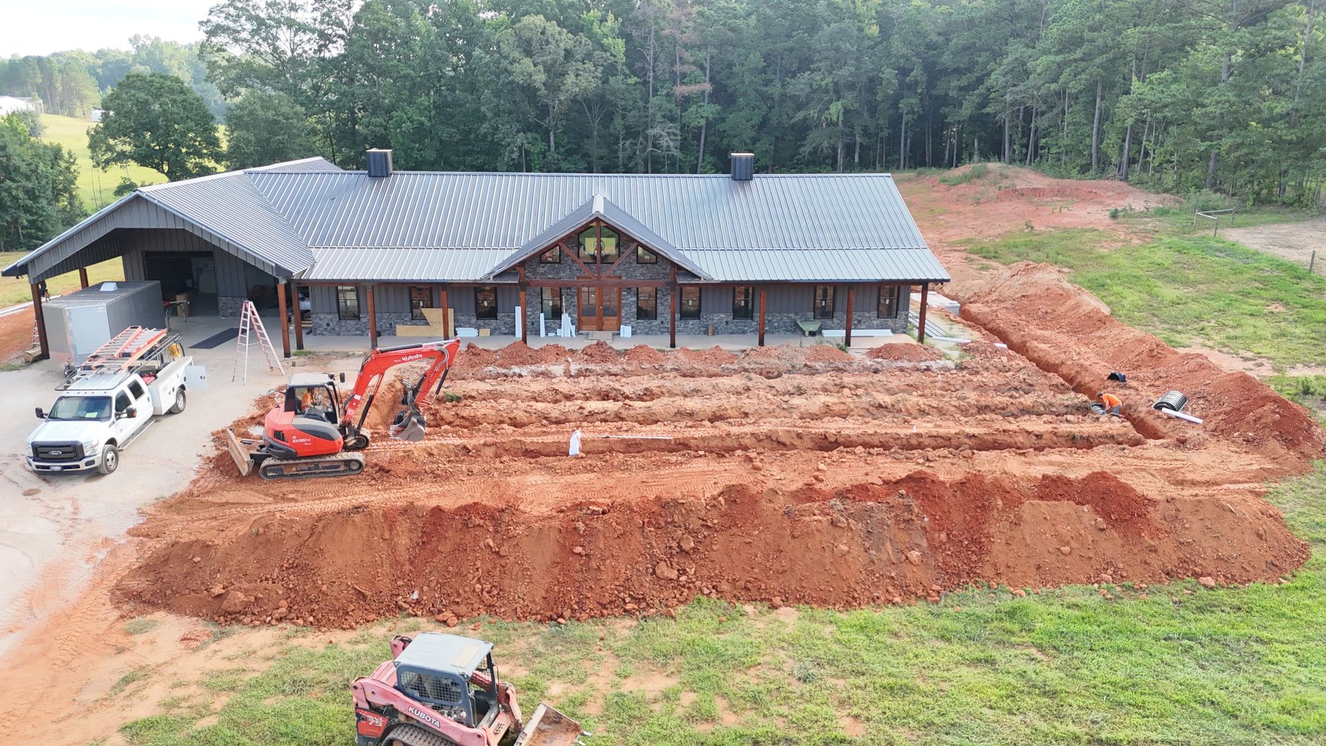Red dirt excavation around a house under construction with an excavator and trucks beside a wooded area.
