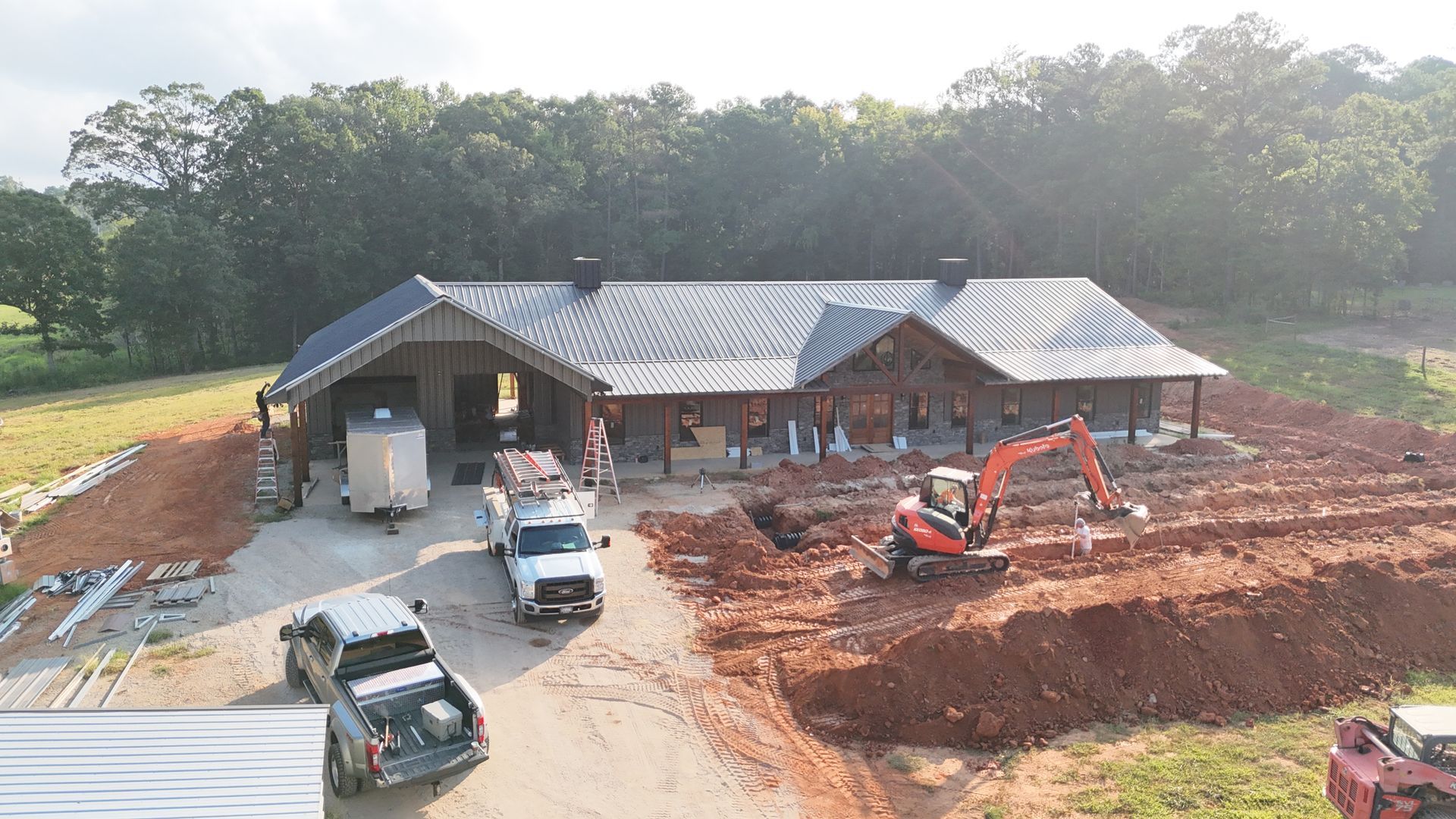 Construction site of a single-story building with a dark roof. An excavator digs, trucks are parked.