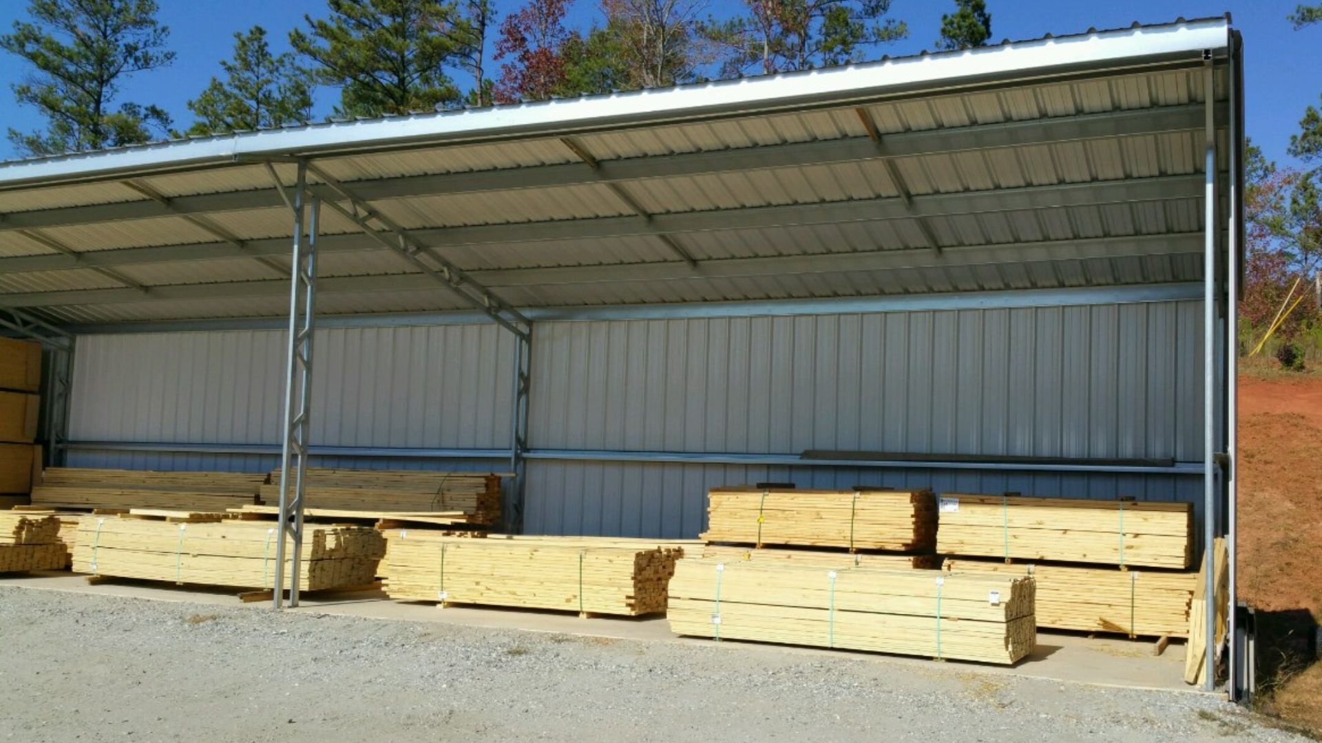 Lumber stacked under a metal roof shed, likely for storage. Exterior shot, sunny day.