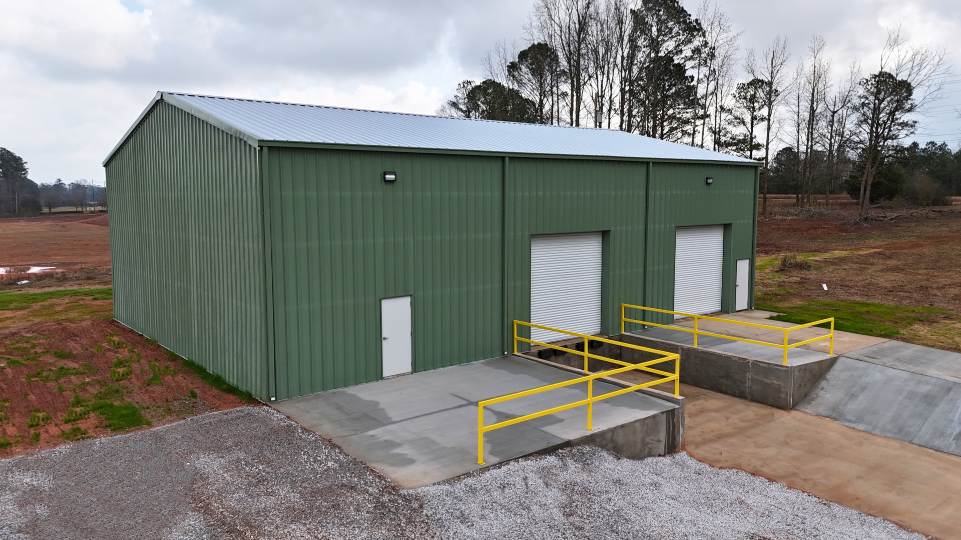 Green metal warehouse with loading docks; gravel area, cloudy sky, trees in the background.