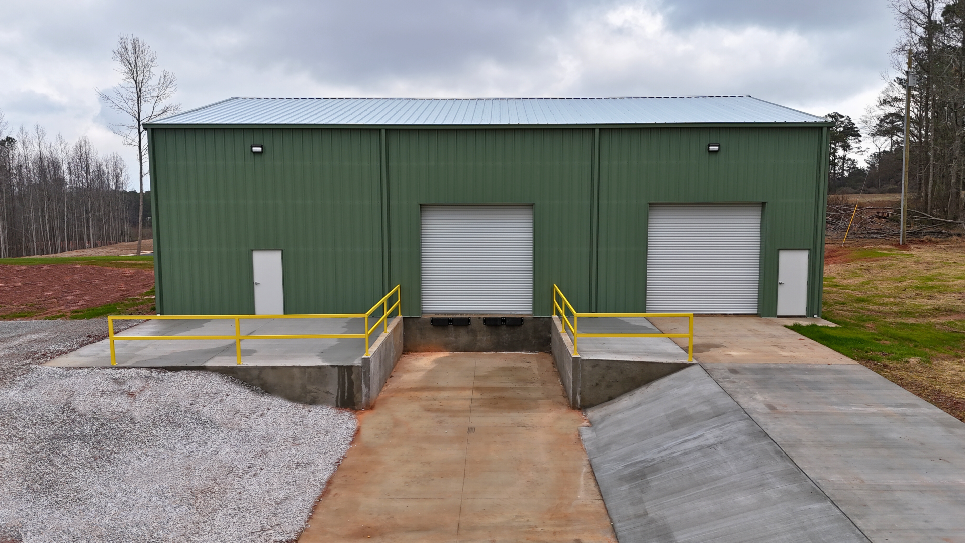 Green metal warehouse with two loading docks, concrete ramp, and gravel.