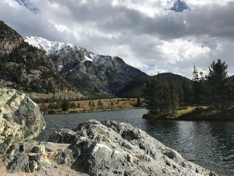 Rocky lakeshore with pine trees, mountains, and cloudy sky reflected on the water at Ripple and Root Therapy in Colorado