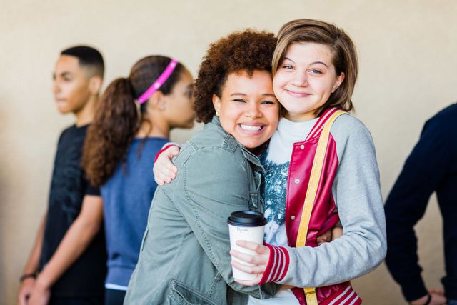 Two young girls are hugging each other while holding a cup of coffee.