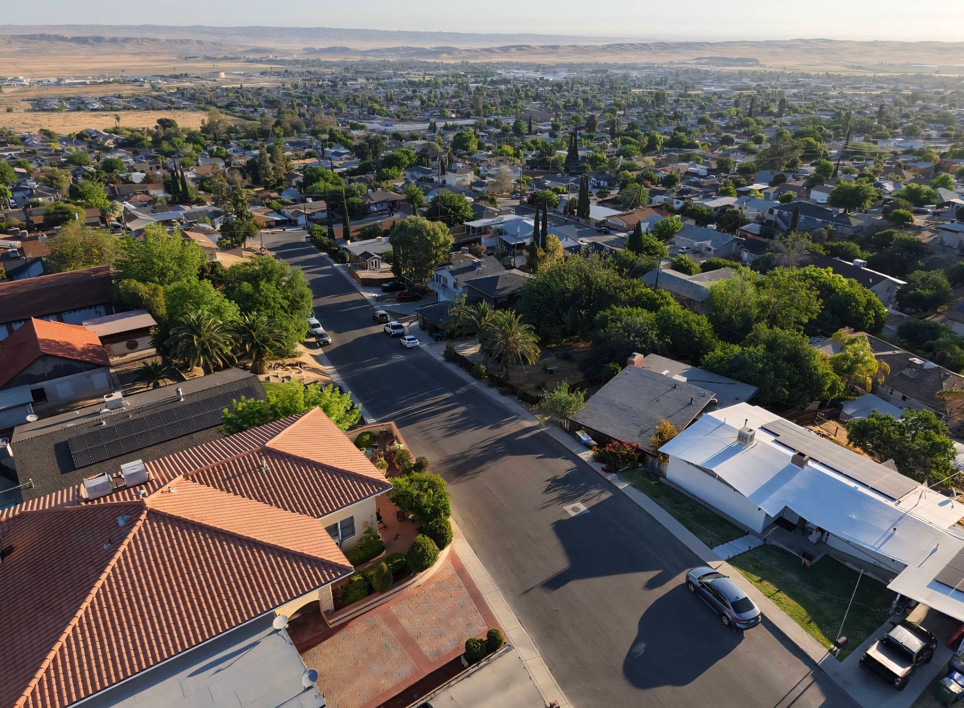 Aerial view of a suburban neighborhood with houses, trees, and streets in warm sunlight.