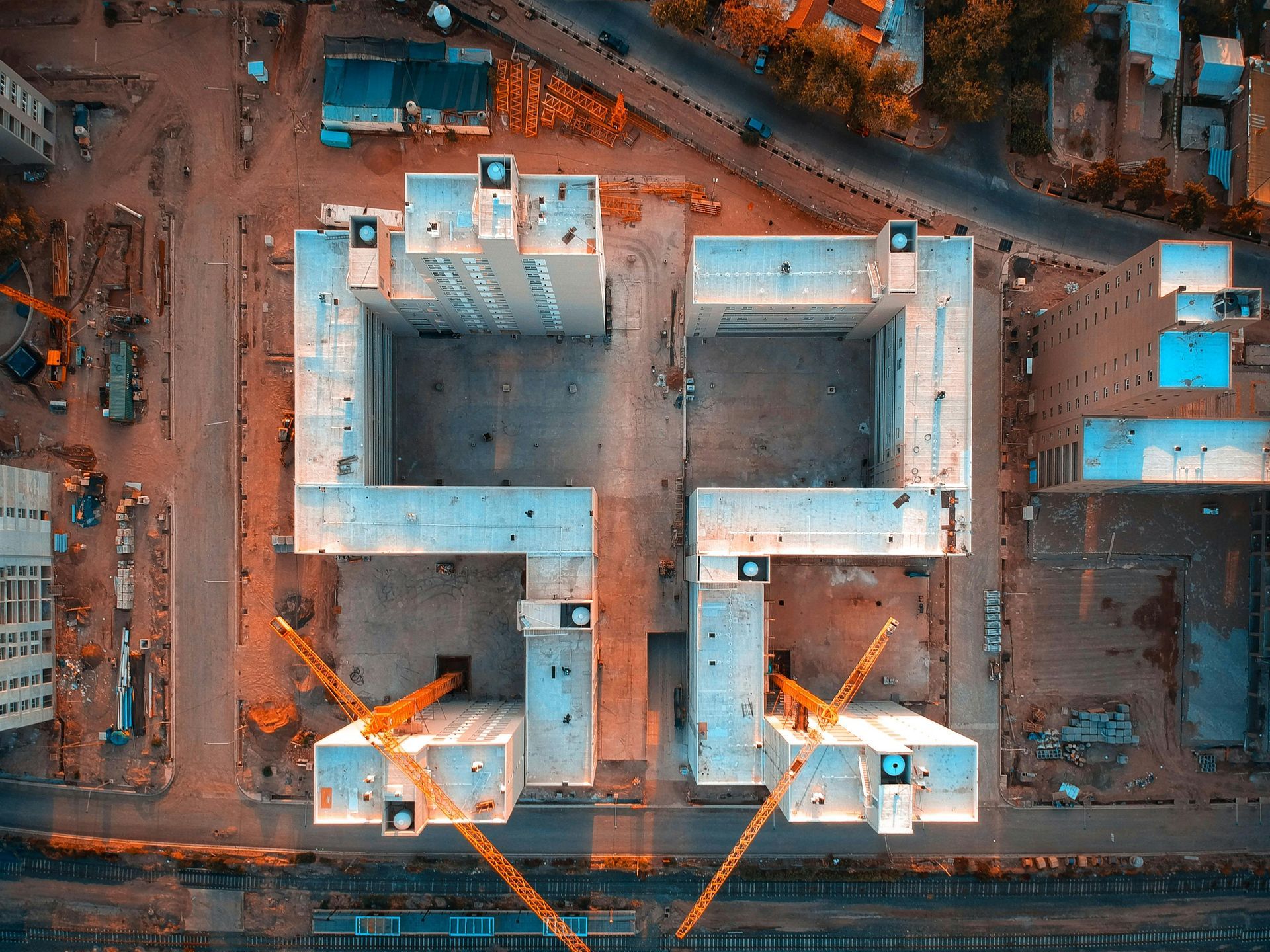 Aerial view of a construction site with two large blue-roofed buildings and orange cranes at dusk