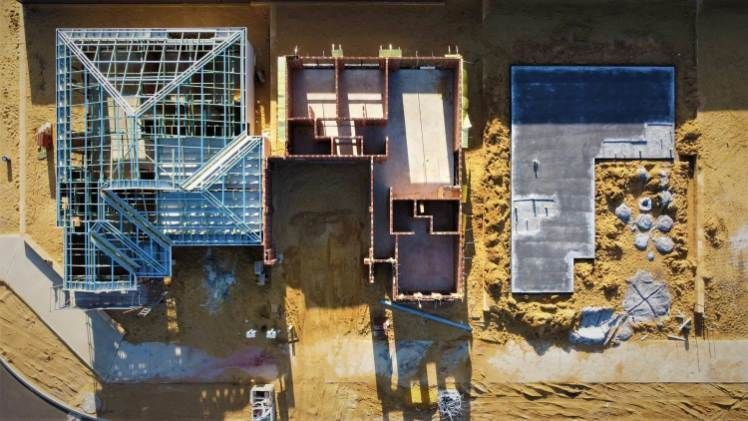 Aerial view of a building construction site with steel framing, roofing materials, and equipment on a dirt lot
