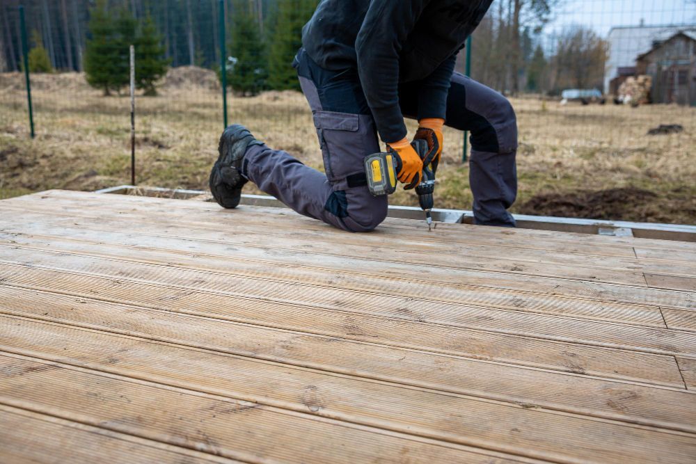 Worker kneeling on wooden deck boards, using a power drill outdoors