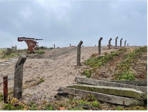 Betonnen trappen en hekpalen op een grasheuvel; een verroest metalen beeldhouwwerk en prikkeldraad zichtbaar aan de bewolkte horizon.