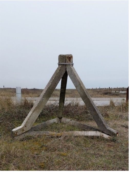 Grijze betonnen driepootconstructie in een grasveld onder een bewolkte hemel.