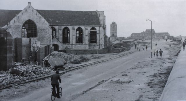 Straatbeeld van door de oorlog beschadigde gebouwen. Een fietser rijdt over de weg, mensen lopen. Een kerk ligt in ruïne.