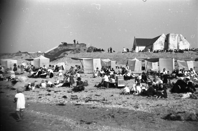 Strandtafereel met veel mensen, tenten en een gebouw op een zandstrand onder een zonnige hemel.