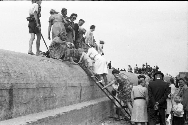 Mensen beklimmen een betonnen constructie met behulp van een ladder, terwijl anderen toekijken. Buiten, mogelijk aan zee.