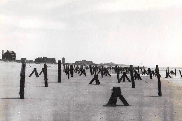 Rijen verweerde houten palen op een zandstrand, met in de verte gebouwen onder een wazige hemel.