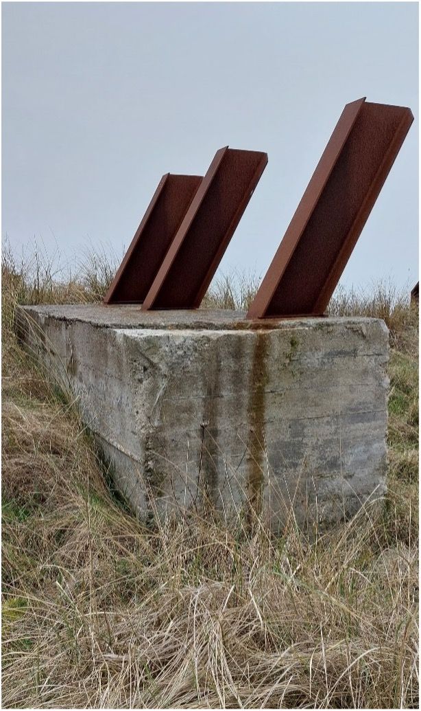 Een betonnen blok met drie roestige metalen balken, geplaatst in hoog, droog gras onder een bewolkte hemel.
