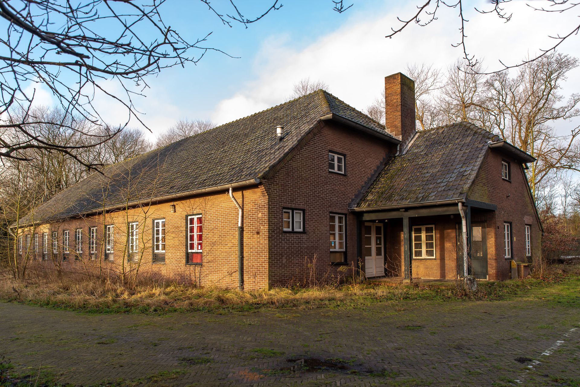 Een bakstenen gebouw met een donker dak en schoorsteen, gelegen tussen kale bomen en verwilderd gras op een zonnige dag.