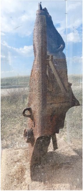 Een verroest metalen voorwerp, mogelijk een machine, staat rechtop in een veld. De lucht is zichtbaar op de achtergrond.