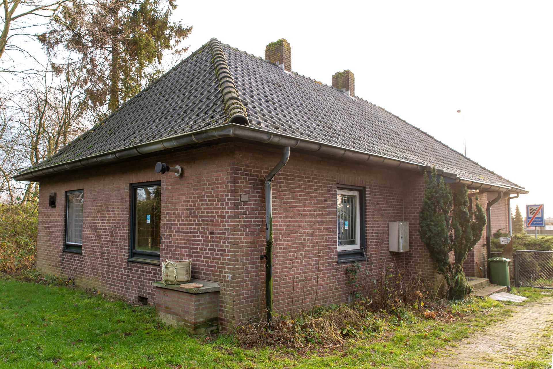 Bakstenen gebouw met een donker pannendak, twee schoorstenen en drie ramen. Gelegen naast een pad en groen.
