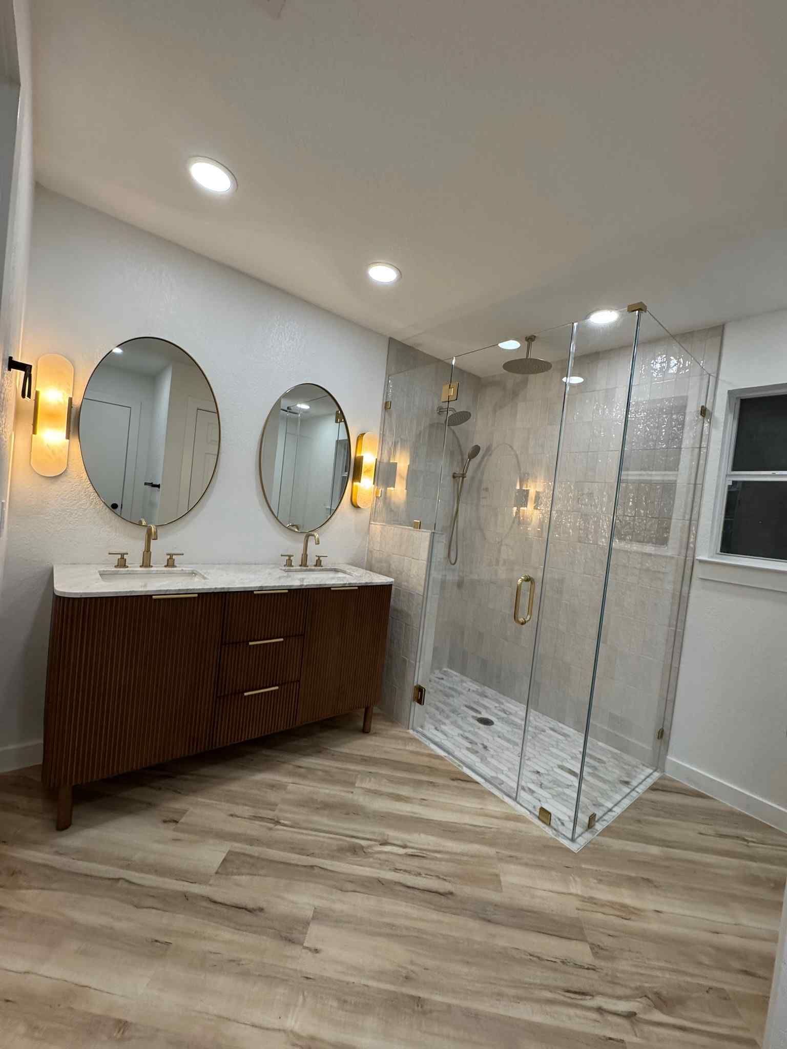 Modern bathroom with double vanity, oval mirrors, and a glass shower on light wood flooring