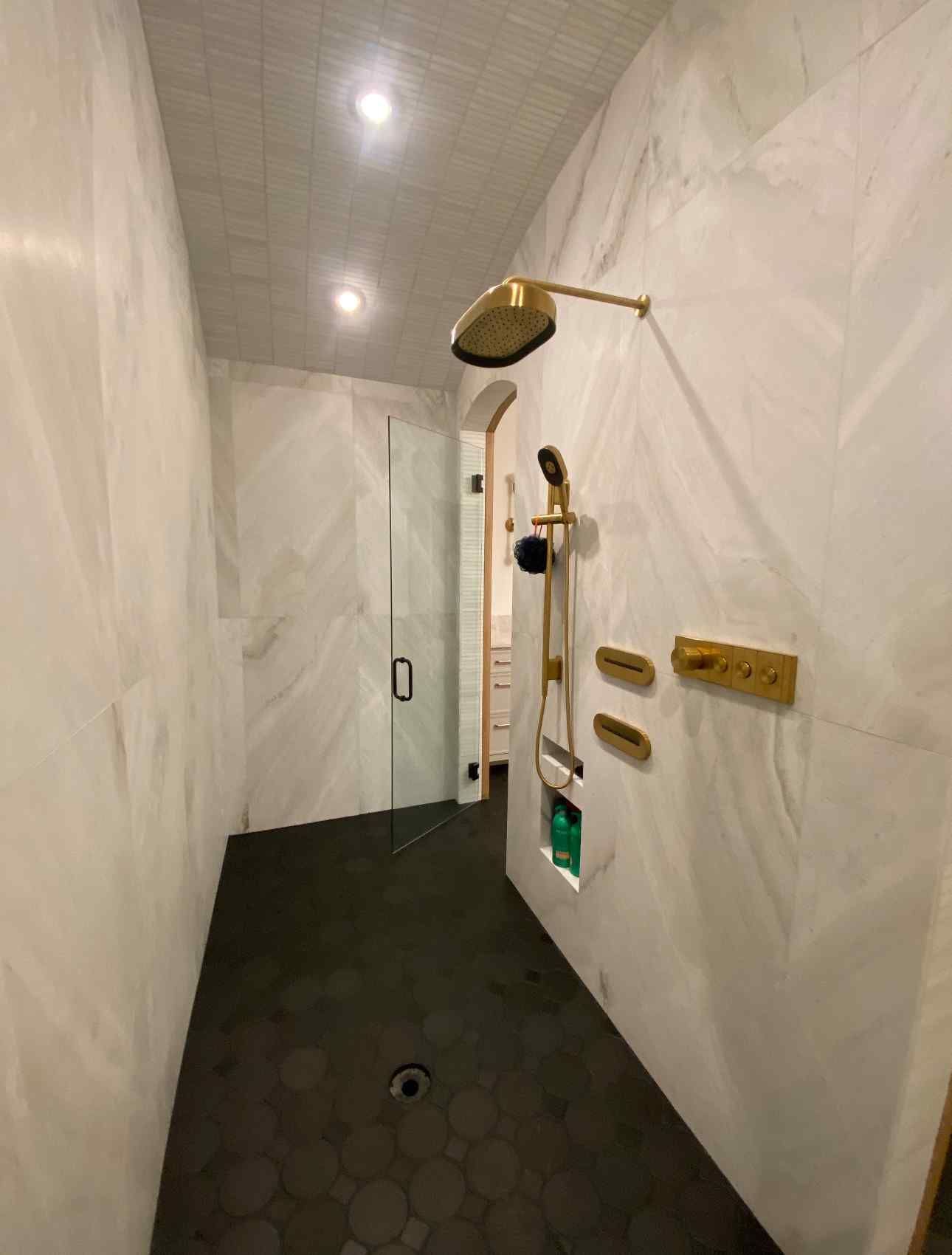 Modern walk-in shower with marble-look walls, black hex tile floor, gold fixtures, and a bench.