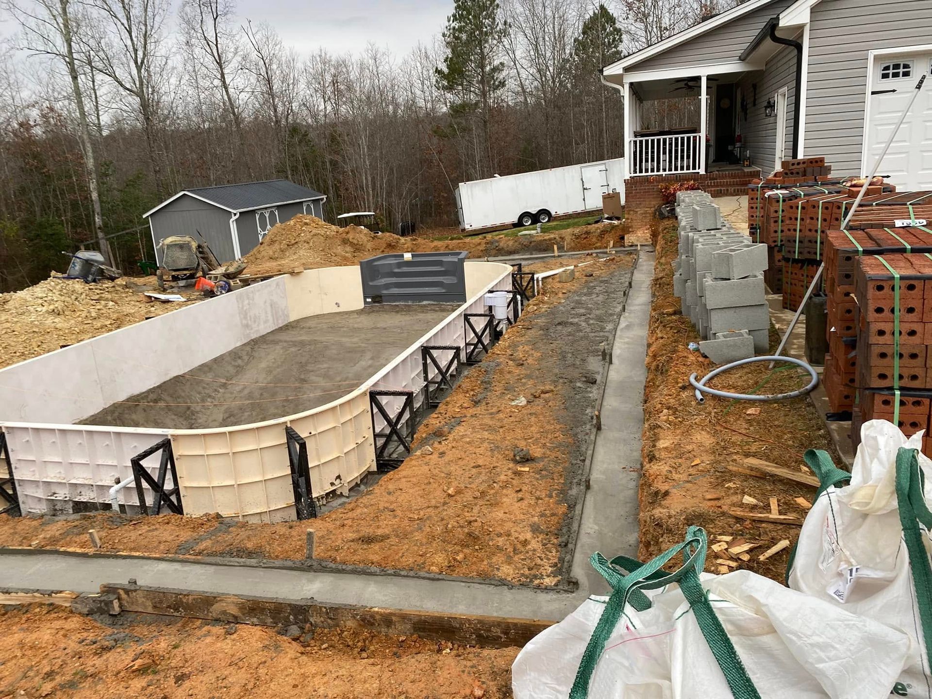 Backyard construction site with an in-ground pool, retaining walls, brick stacks, and a house porch.