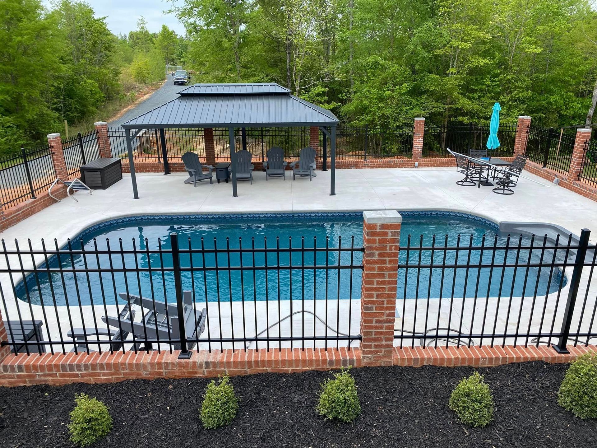 Backyard pool with black fence, brick patio, pergola, lounge chairs, and trees beyond