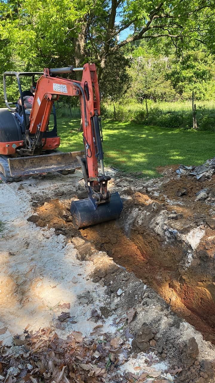 Orange excavator digging a muddy trench beside a grassy, tree-lined area.