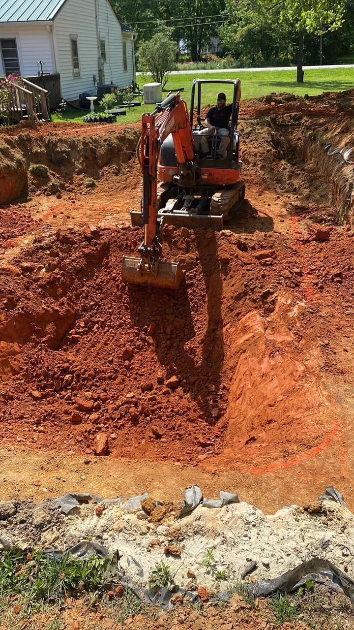 Excavator digging a deep red-soil pit beside a house on a sunny day