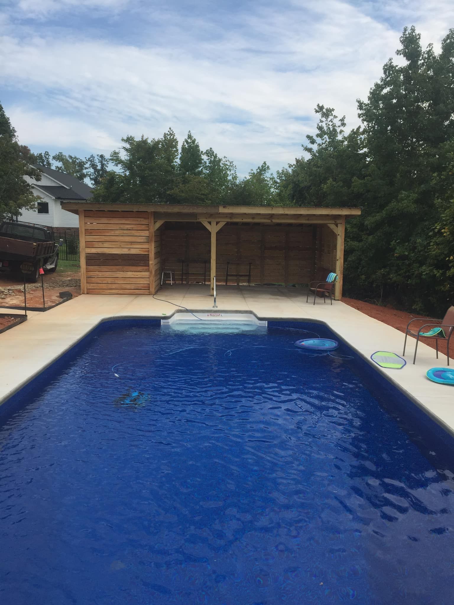 Backyard swimming pool with a wooden cabana and trees under a cloudy sky