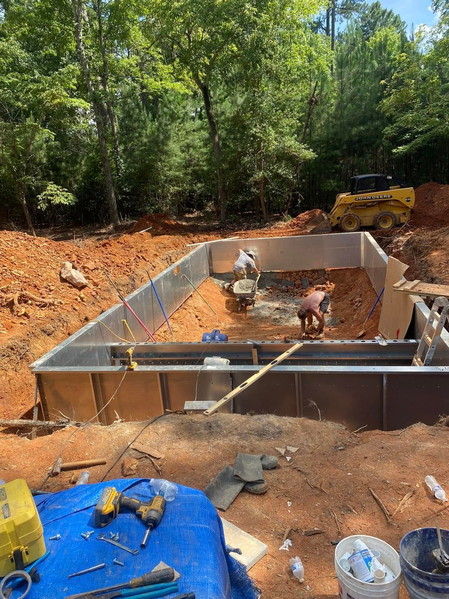 Rectangular concrete foundation at a wooded construction site, with dirt, tools, and blue tarp in the foreground