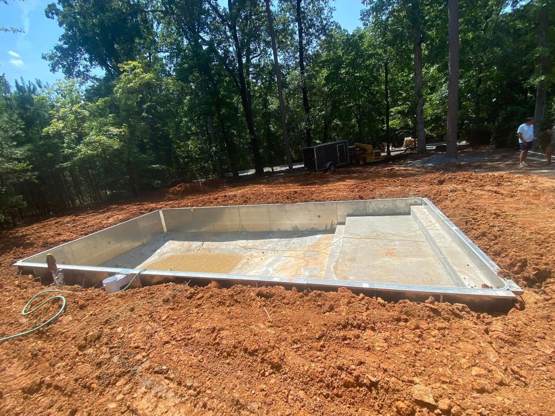 Empty rectangular pool shell in a wooded yard, surrounded by reddish dirt and trees.