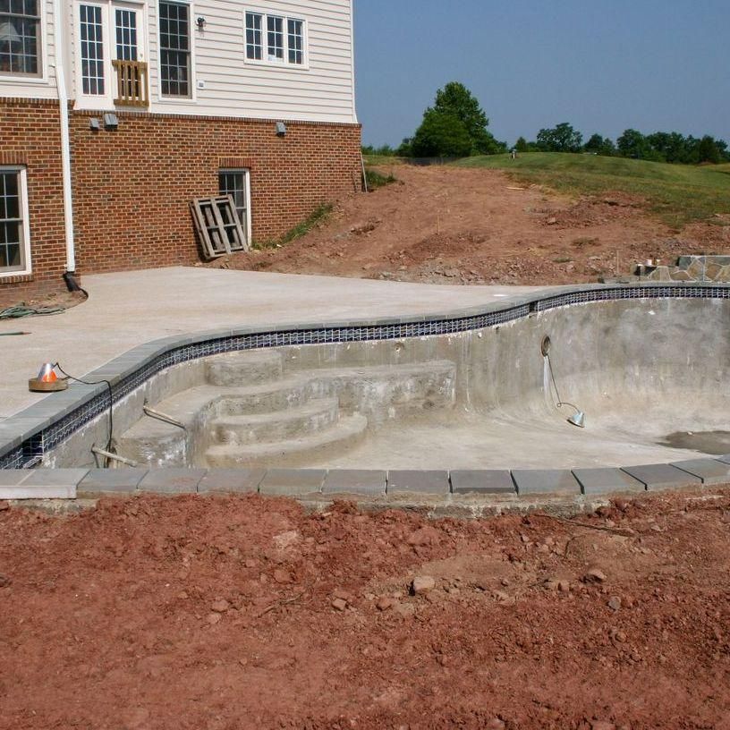 Empty in-ground pool beside a house, with unfinished red dirt and concrete steps.