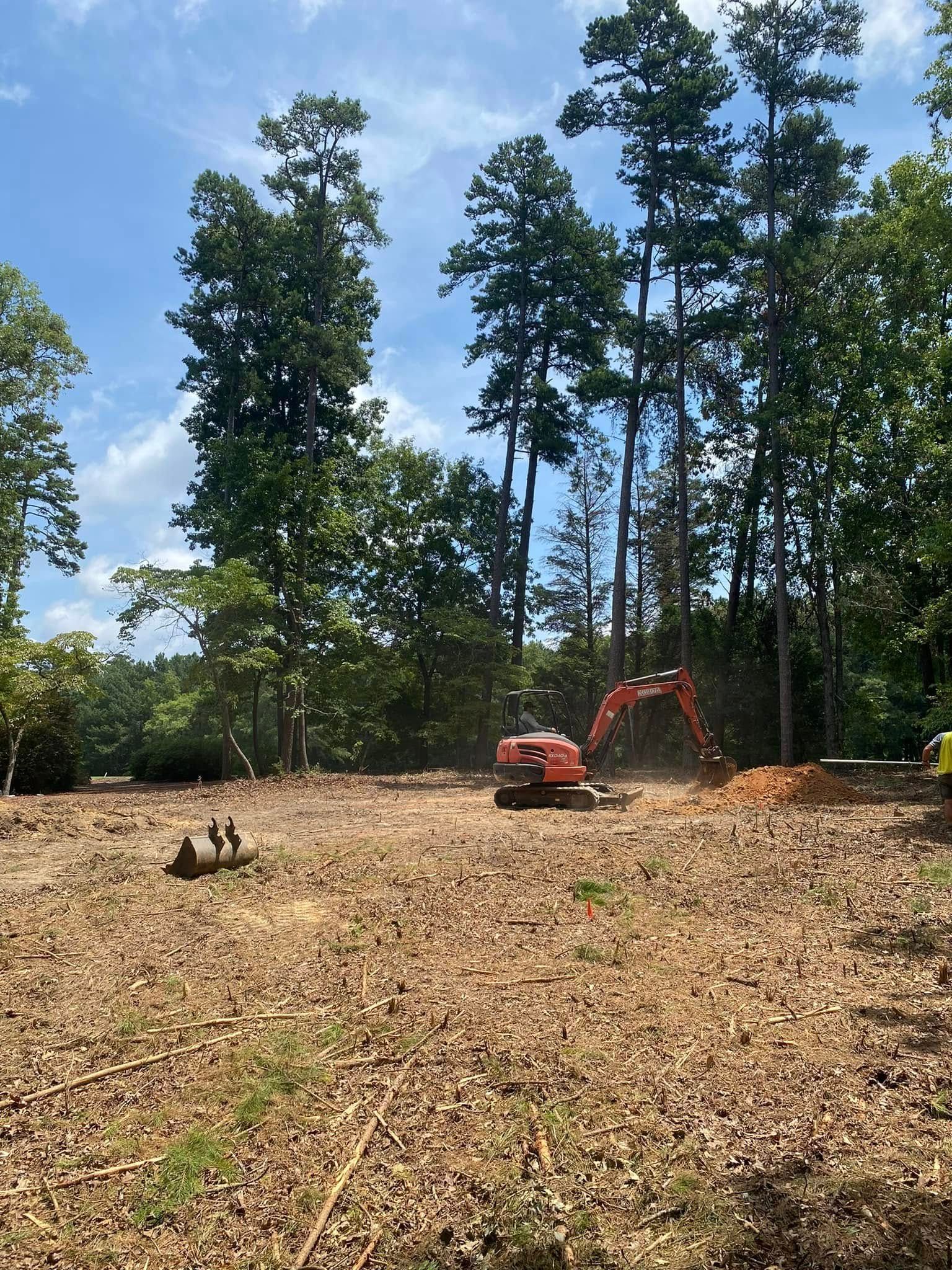 Excavator and small orange truck on a cleared dirt lot with tall trees under a blue sky