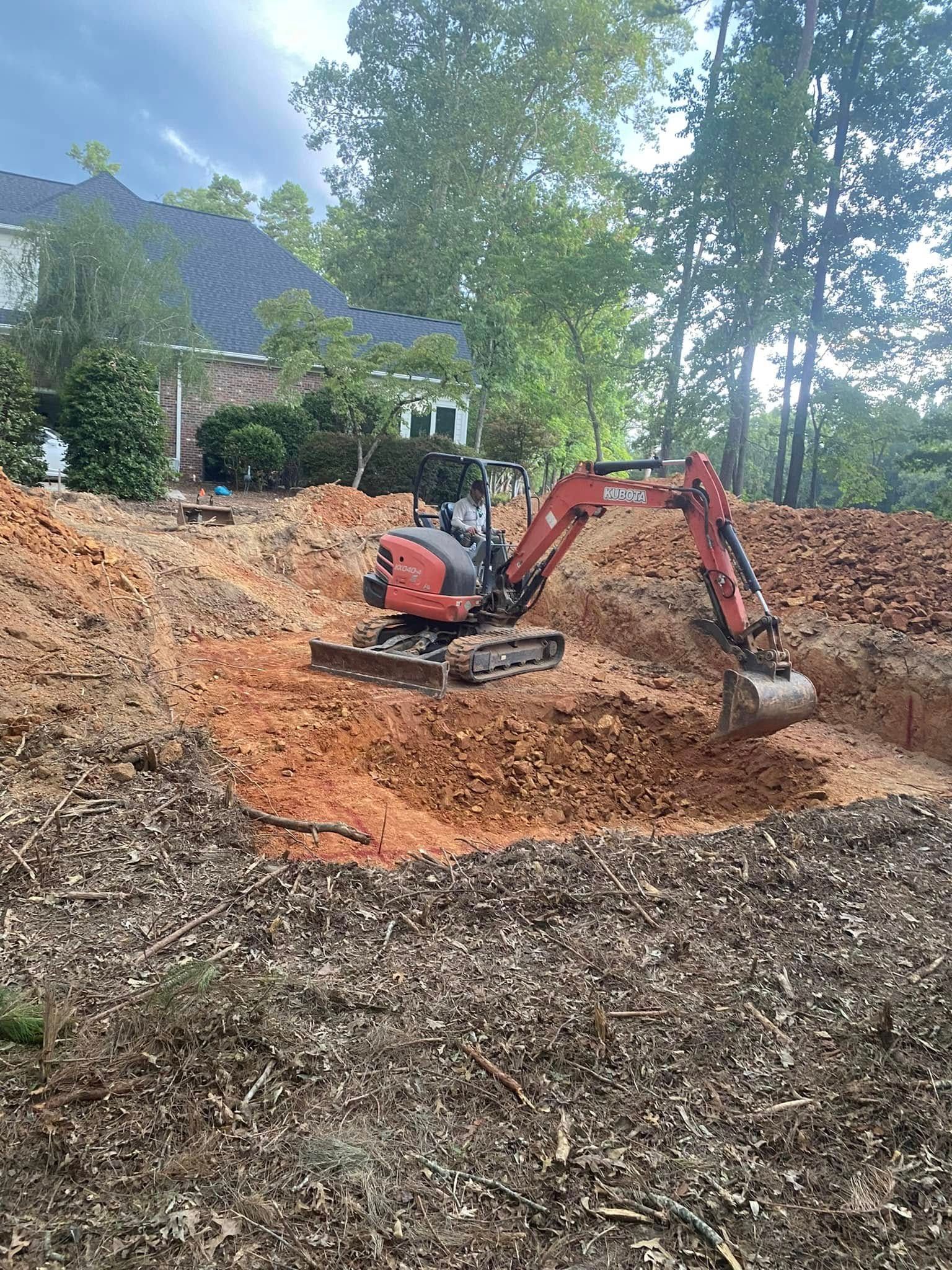 Orange mini excavator digging in a dirt lot beside trees and a house under construction