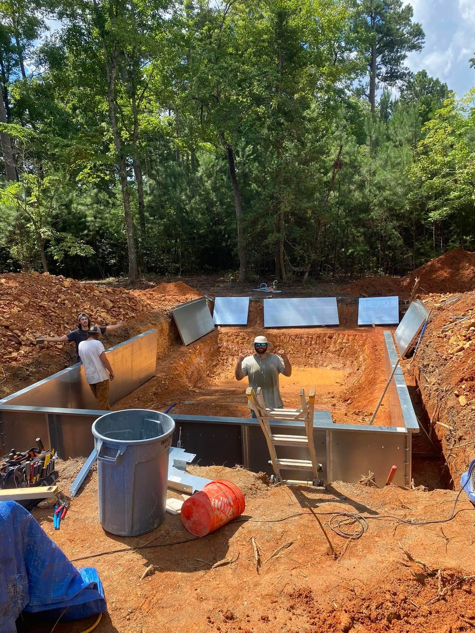Workers building a concrete pit in a wooded area, with steel forms and orange dirt around the site.