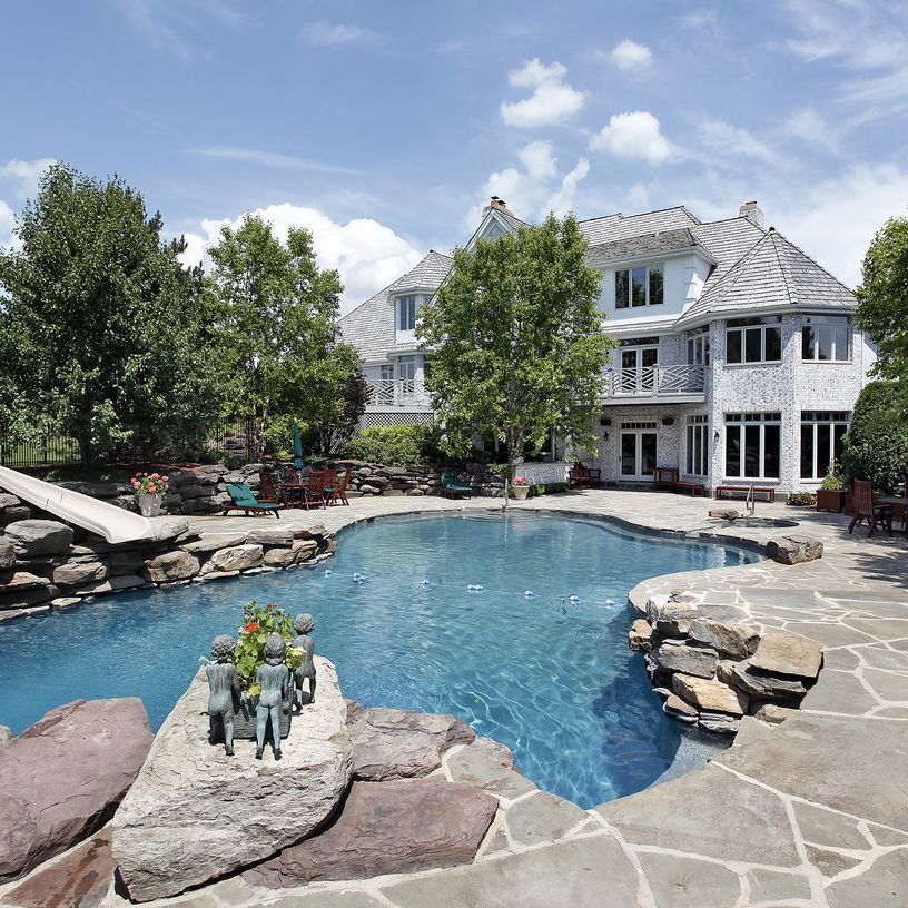 Backyard pool with stone patio, rock landscaping, and a large gray house under a blue sky