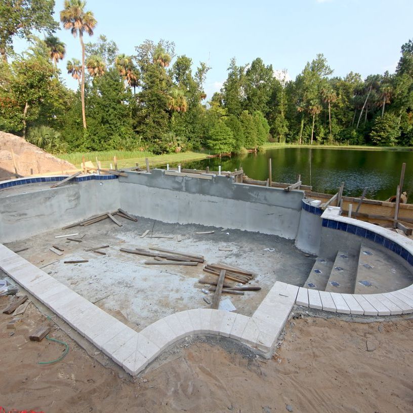 Empty backyard swimming pool under construction beside a pond, with trees in the background