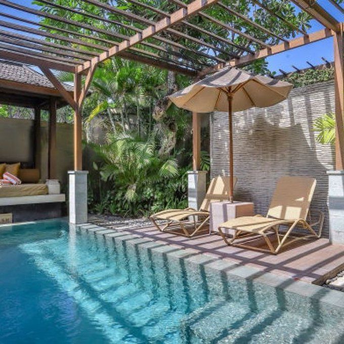 Modern patio with a narrow blue pool, two lounge chairs, umbrella, and lush tropical plants