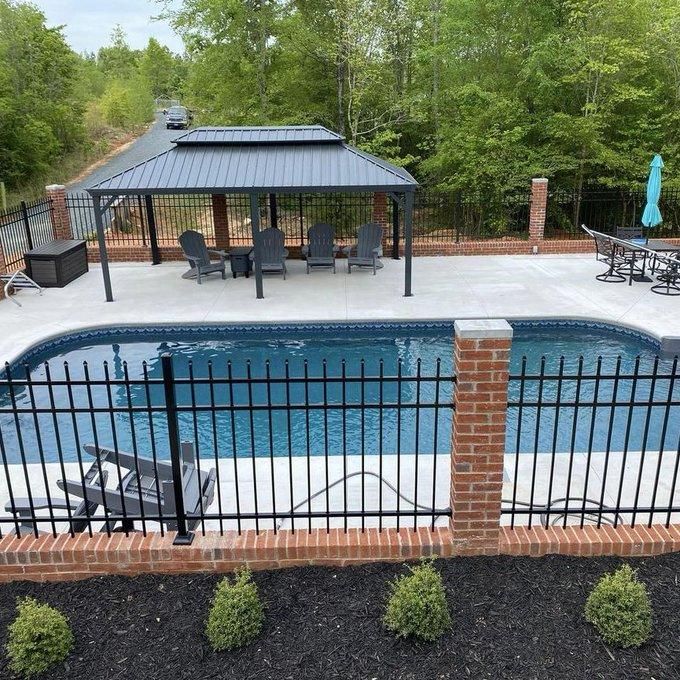 Backyard pool with black fence, covered patio seating, brick columns, and surrounding trees