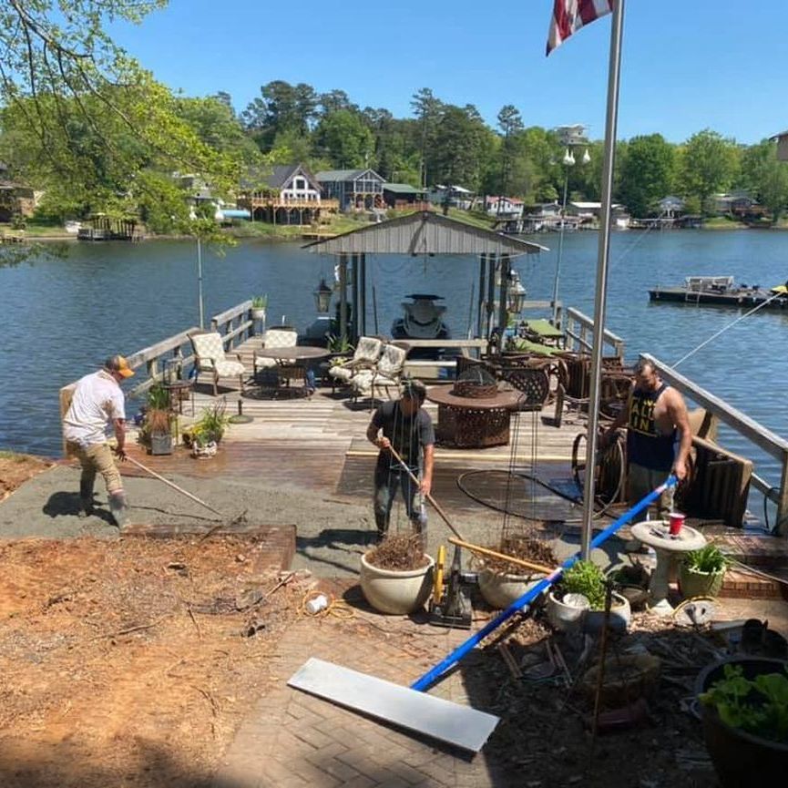 Workers planting along a lakeside dock with potted plants and a flag in the background