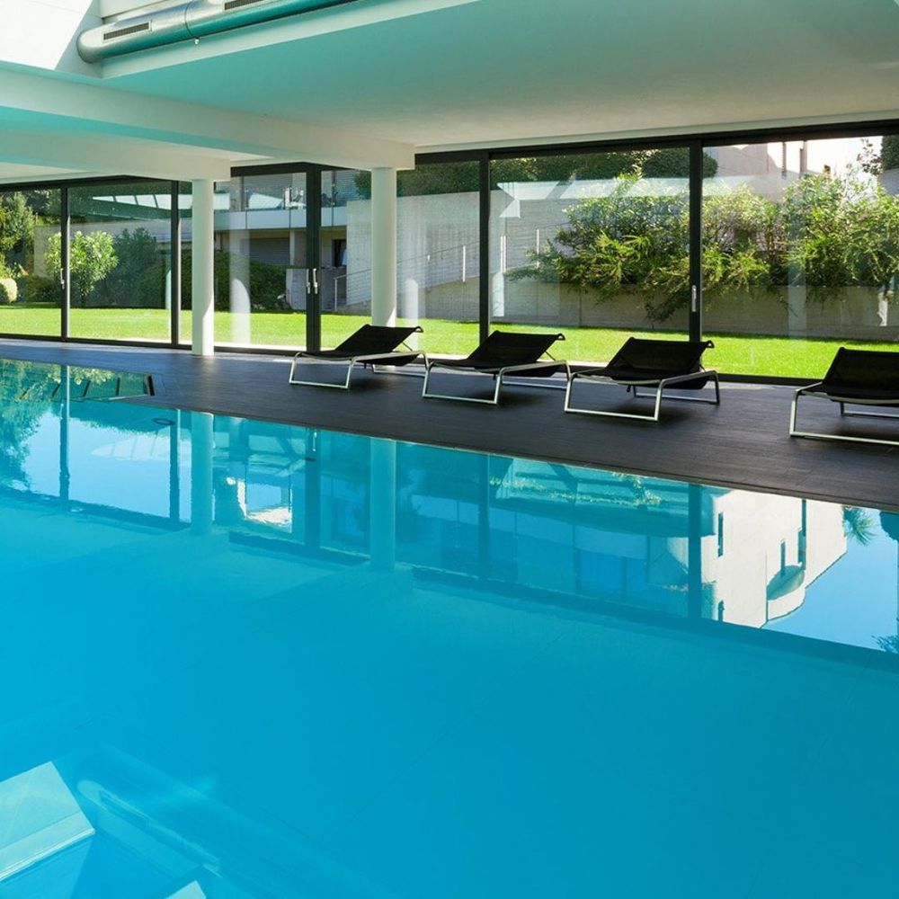 Modern indoor pool with turquoise water, lounge chairs, and large glass walls overlooking a green yard