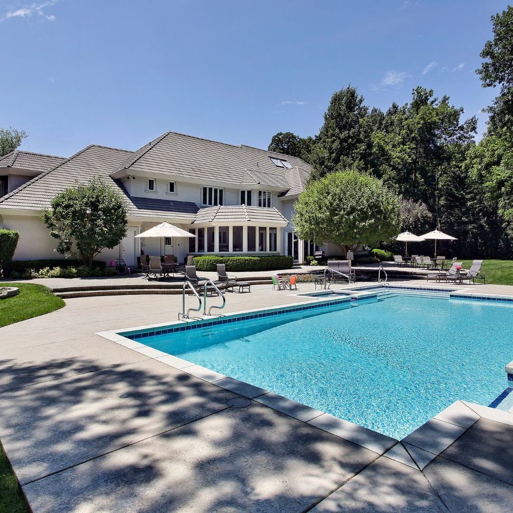 Backyard swimming pool beside a two-story house with lounge chairs and patio umbrellas on a sunny day