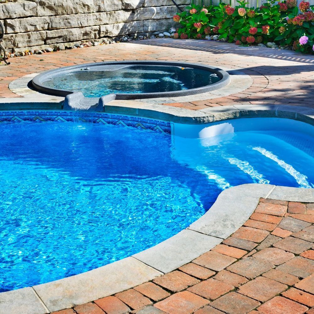 Blue backyard pool with a built-in hot tub and stone patio, bordered by flowers and a brick walkway