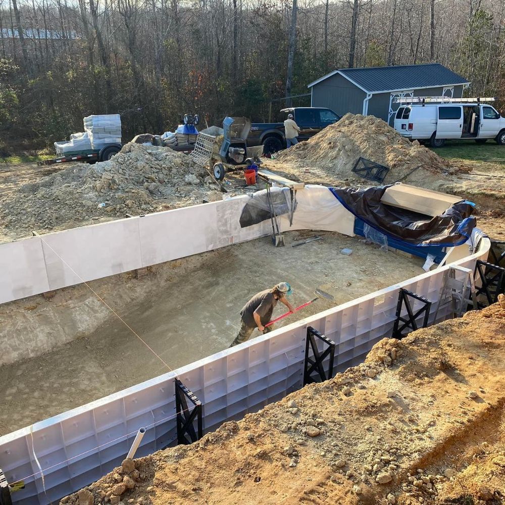 Construction site with a large rectangular pool foundation, workers, gravel piles, and vehicles nearby.