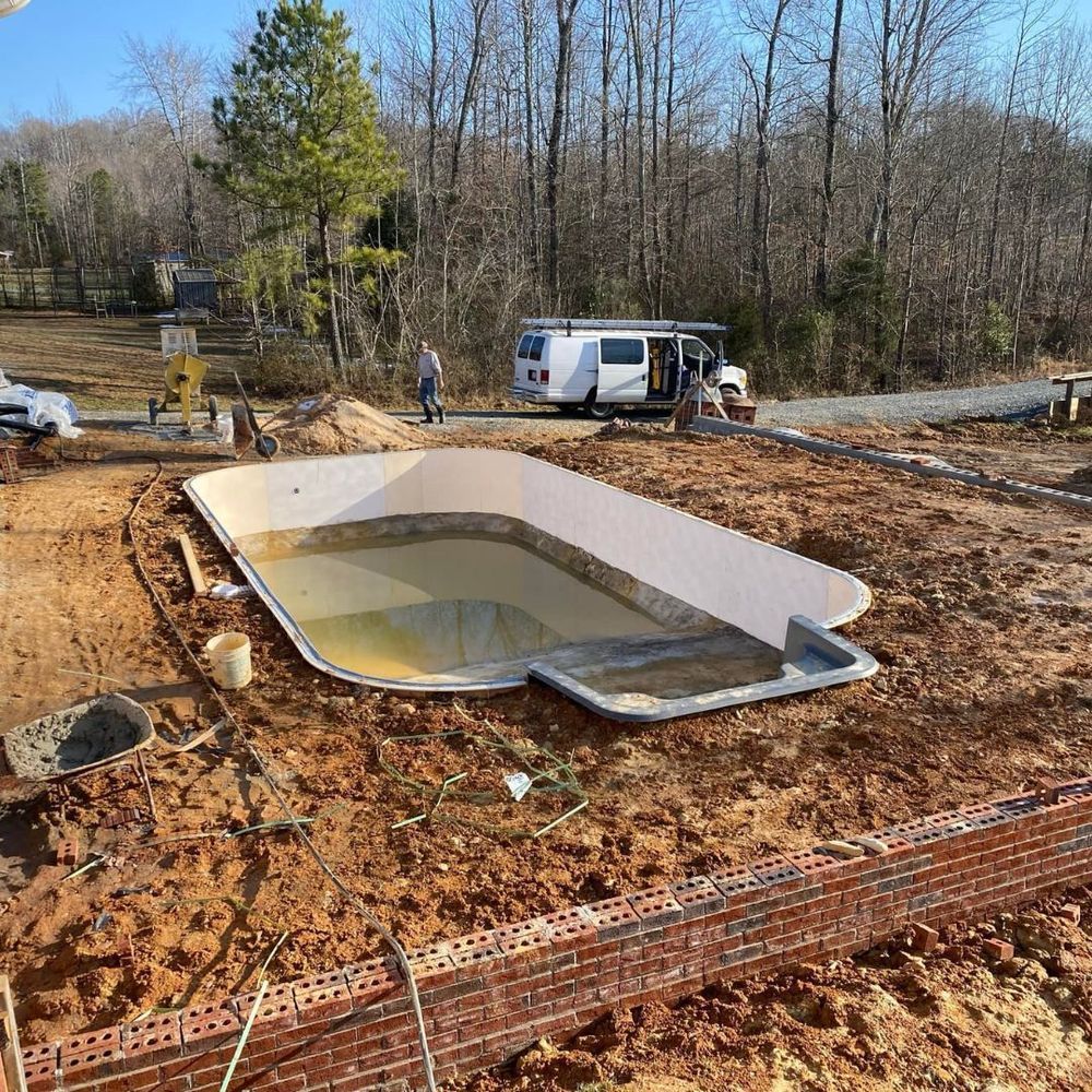 Partially built backyard pool with water, surrounded by dirt and construction materials near a wooded area