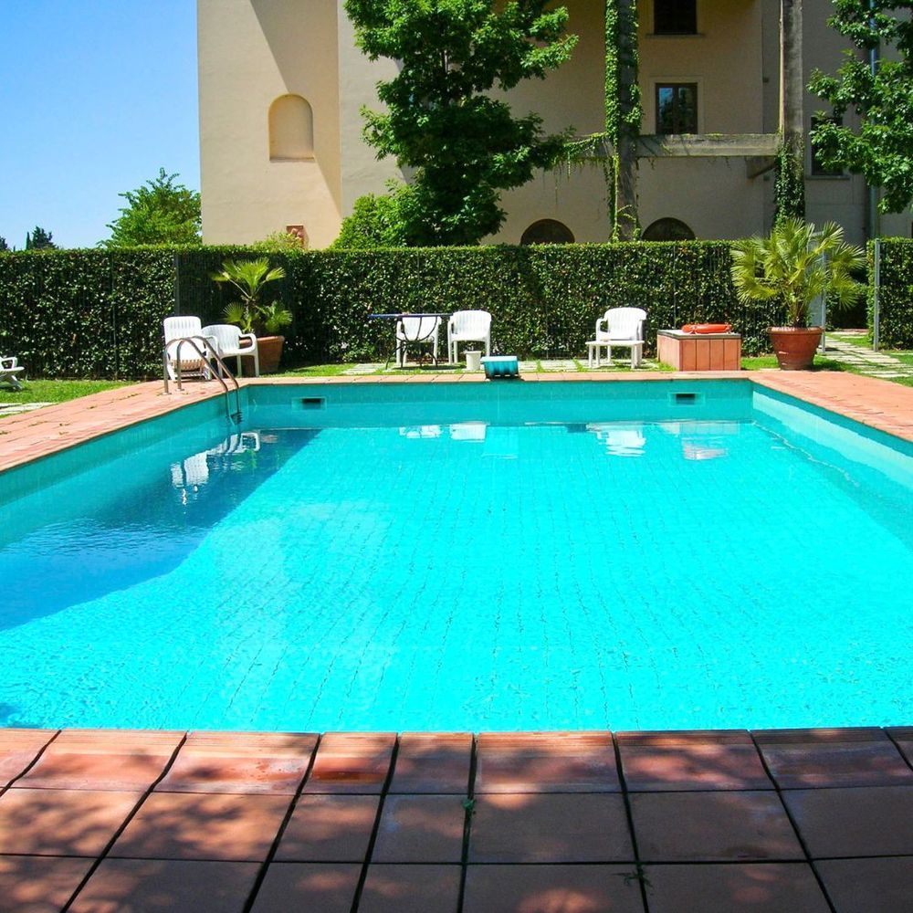 Blue swimming pool beside a beige building, with white chairs and greenery under bright sunlight