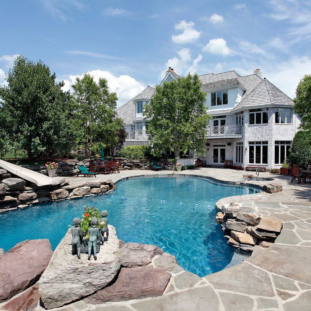 Backyard pool beside a large white house with stone patio and trees under a blue sky